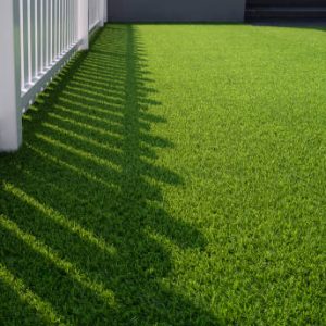 Sunlight and shadow of white wooden fence on green artificial turf surface in front yard of home, selective focus with high angle view